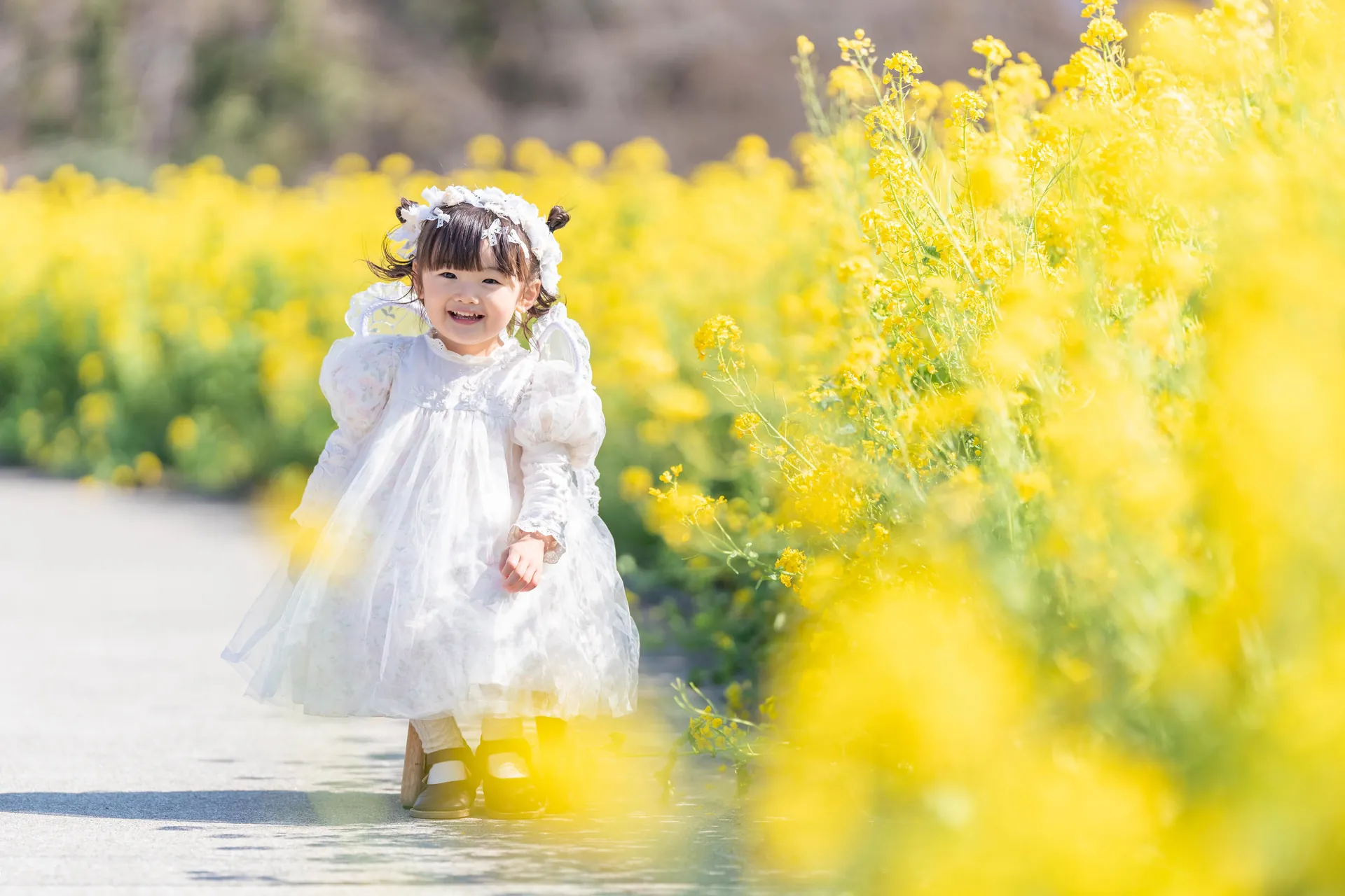 菜の花畑の小道で、白いドレスの子どもが笑顔を見せている写真